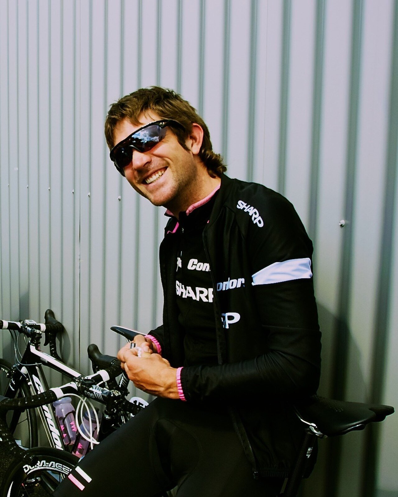 Dan Craven souriant, lunettes de soleil, appuyé contre un mur à côté des vélos