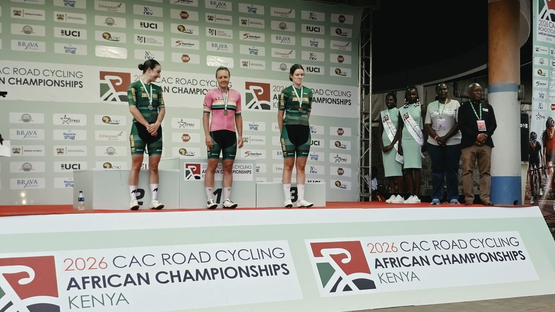 The three South Africans on the podium of the elite women's road race of the 2026 African Road Championships in Kisumu, their medals around their necks, the winner in a pink jersey in the centre; officials and hostesses on the right