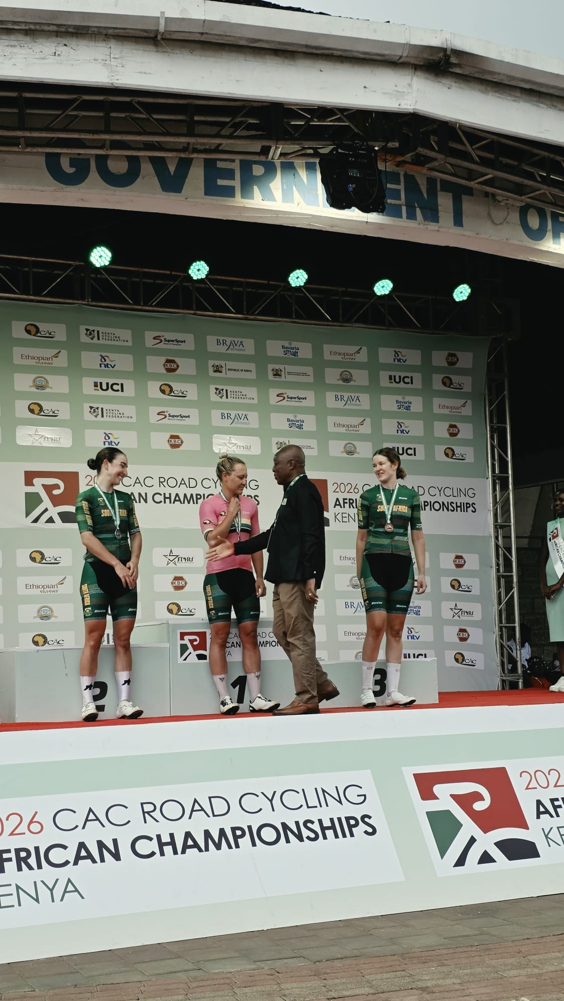 An official shakes hands with the winner in a pink jersey on the top step of the podium, between the second and the third, all in South Africa jerseys