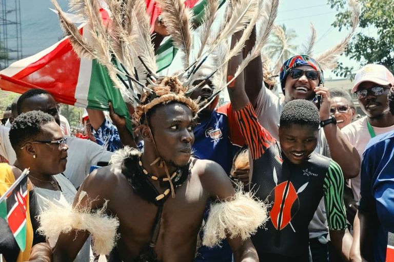 A dancer in a feathered traditional costume, in a crowd waving the flags of Kenya, next to a smiling rider of the Kenyan team