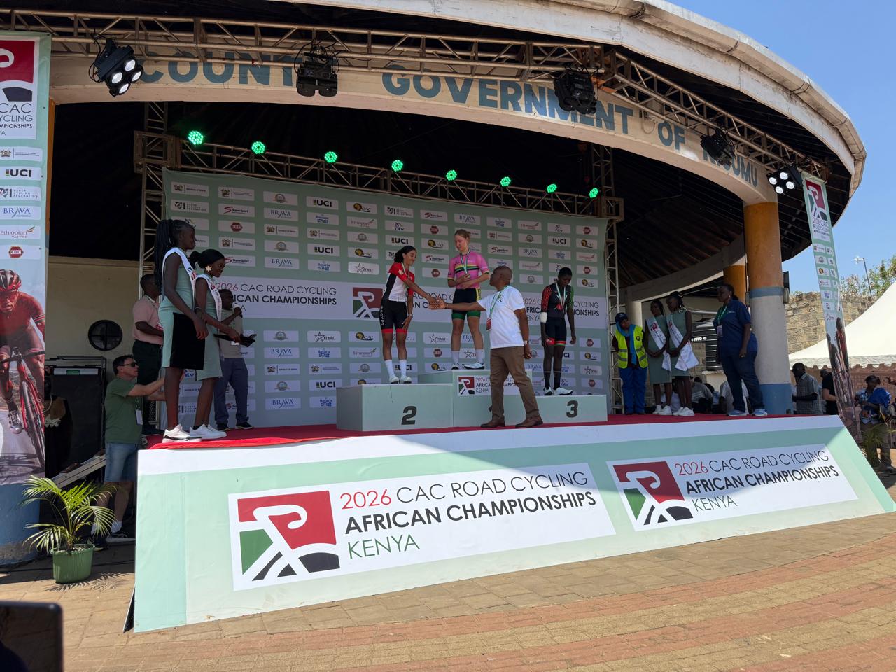 The first three of the junior women's road race on the podium of the 2026 African Road Championships in Kisumu; an official shakes hands with the runner-up