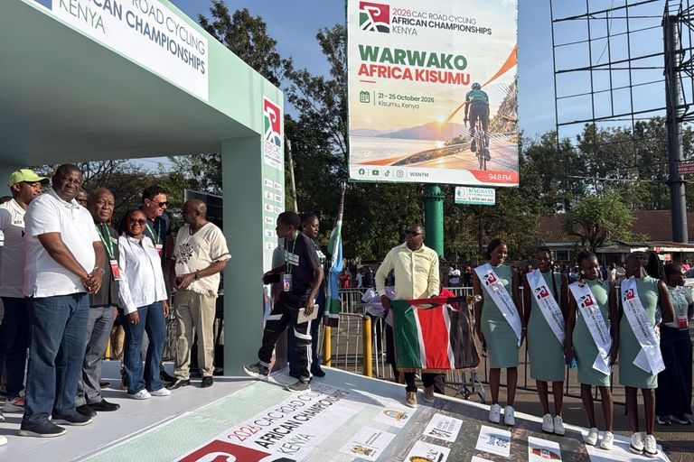 Officials on the start podium of the 2026 African Road Championships in Kisumu, next to a man holding the flag of Kenya and the championships' hostesses, under the championships' billboard