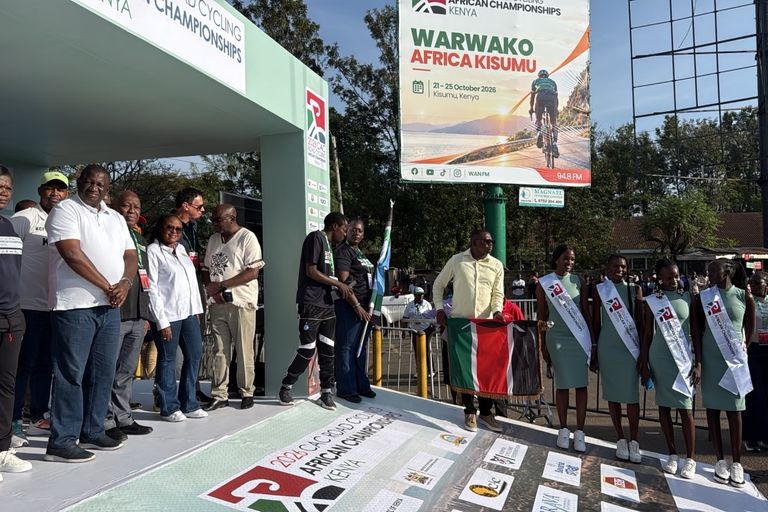 Officials on the start podium of the 2026 African Road Championships in Kisumu; in front of them, a man holds the flag of Kenya, under the championships' billboard