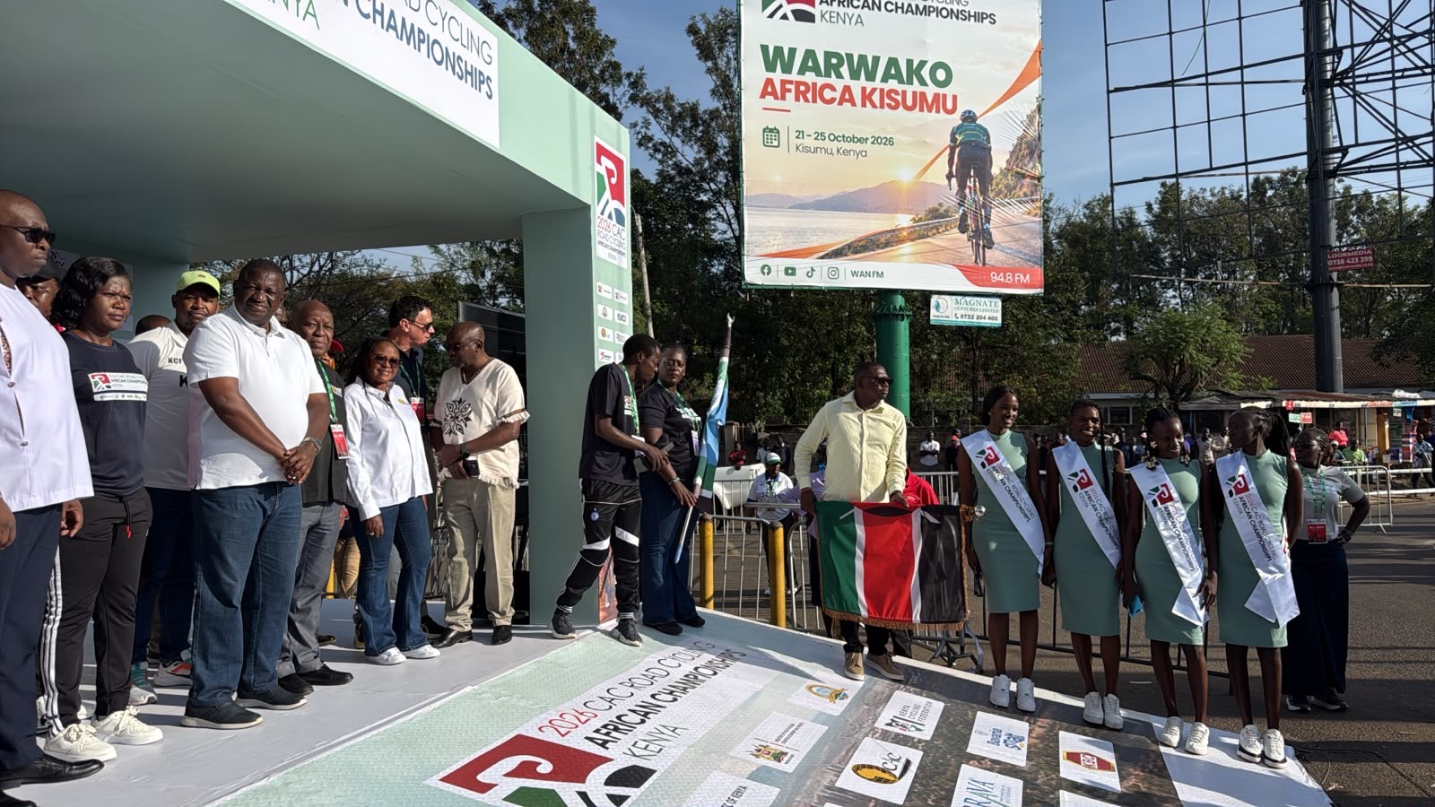 Officials on the start podium of the 2026 African Road Championships in Kisumu; in front of them, a man holds the flag of Kenya, under the championships' billboard