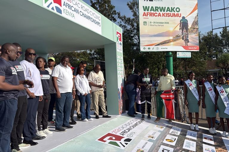 Officials on the start podium of the 2026 African Road Championships in Kisumu, next to the flag of Kenya and the championships' hostesses