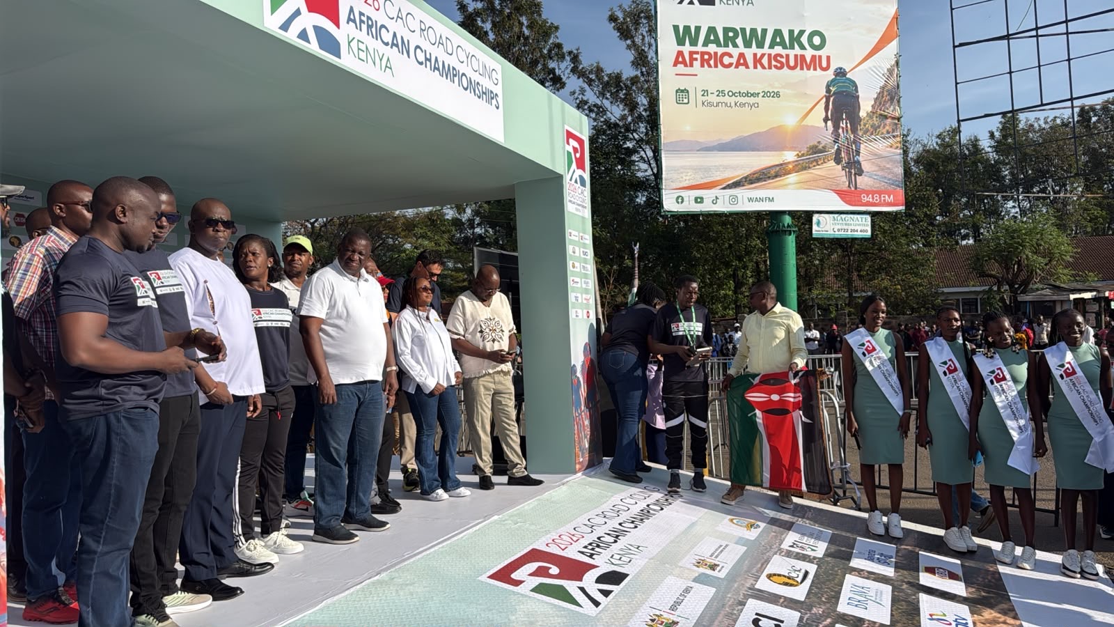 Des officiels sur le podium de départ des championnats d'Afrique sur route 2026 à Kisumu, à côté du drapeau du Kenya et des hôtesses des championnats